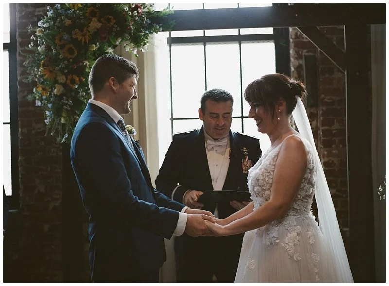A bride and groom holding hands during their wedding ceremony, with an officiant standing behind them in a rustic indoor setting with brick walls and a large window, adorned with floral arrangements.
