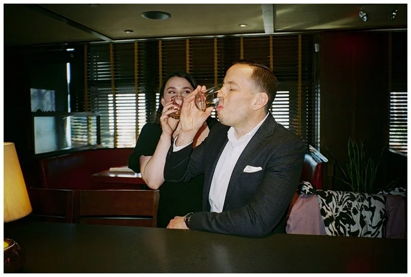 A man in a suit and a woman in a black dress sharing drinks inside a dimly lit restaurant or bar.