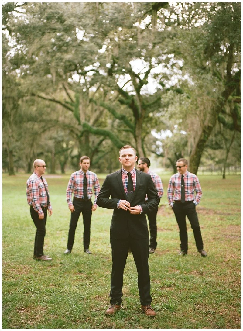 A man in a black suit and plaid shirt standing in front of four men in plaid shirts and sunglasses, outdoors in a park with large trees and green grass.