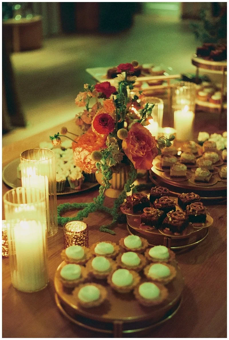 A table decorated with lit candles, and topped with a floral arrangement of orange and pink flowers, surrounded by various desserts including cookies with white frosting, chocolate squares with nuts, and other sweet treats in a dim, warm-lit setting.