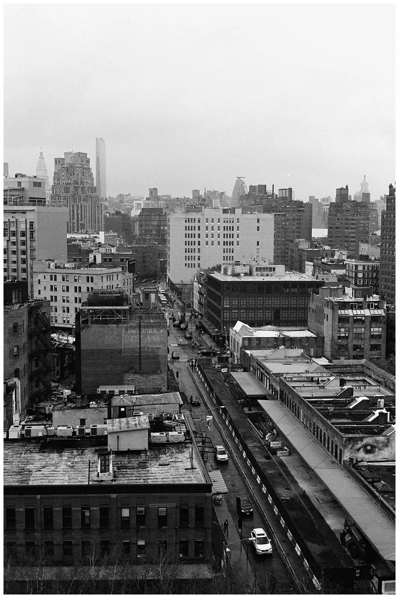 Black and white photo of a city skyline showing multiple tall buildings, some under construction, with streets and parked cars in the foreground.