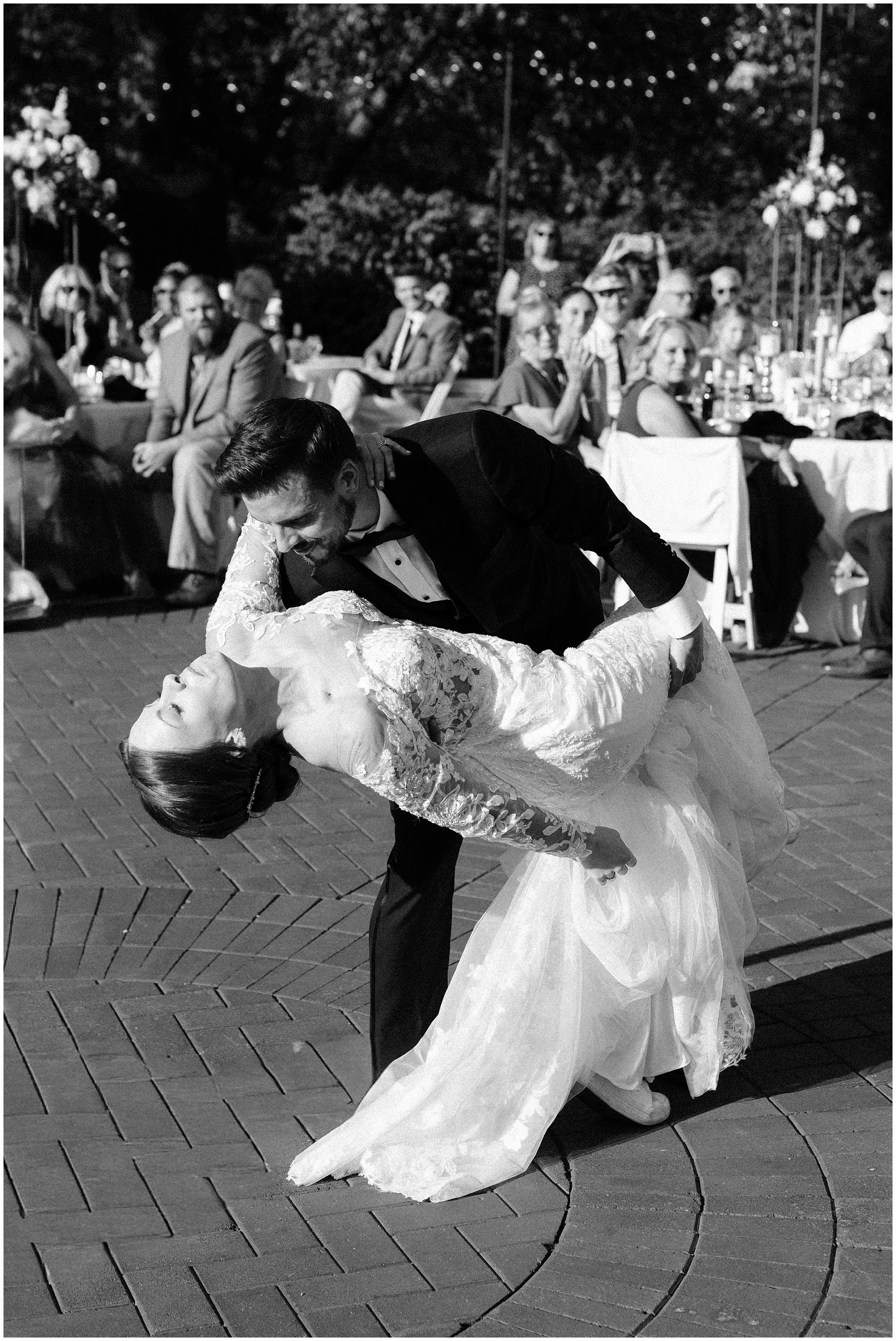 A black-and-white photo of a bride and groom dancing, with the groom dipping the bride. Guests are sitting and watching in the background at an outdoor wedding reception. Franklin Park Conservatory, outdoor wedding, Adam lowe photography