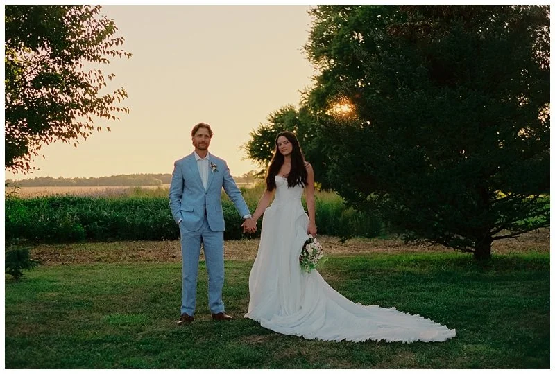A newlywed couple holding hands outdoors during sunset, with trees and an open field in the background.