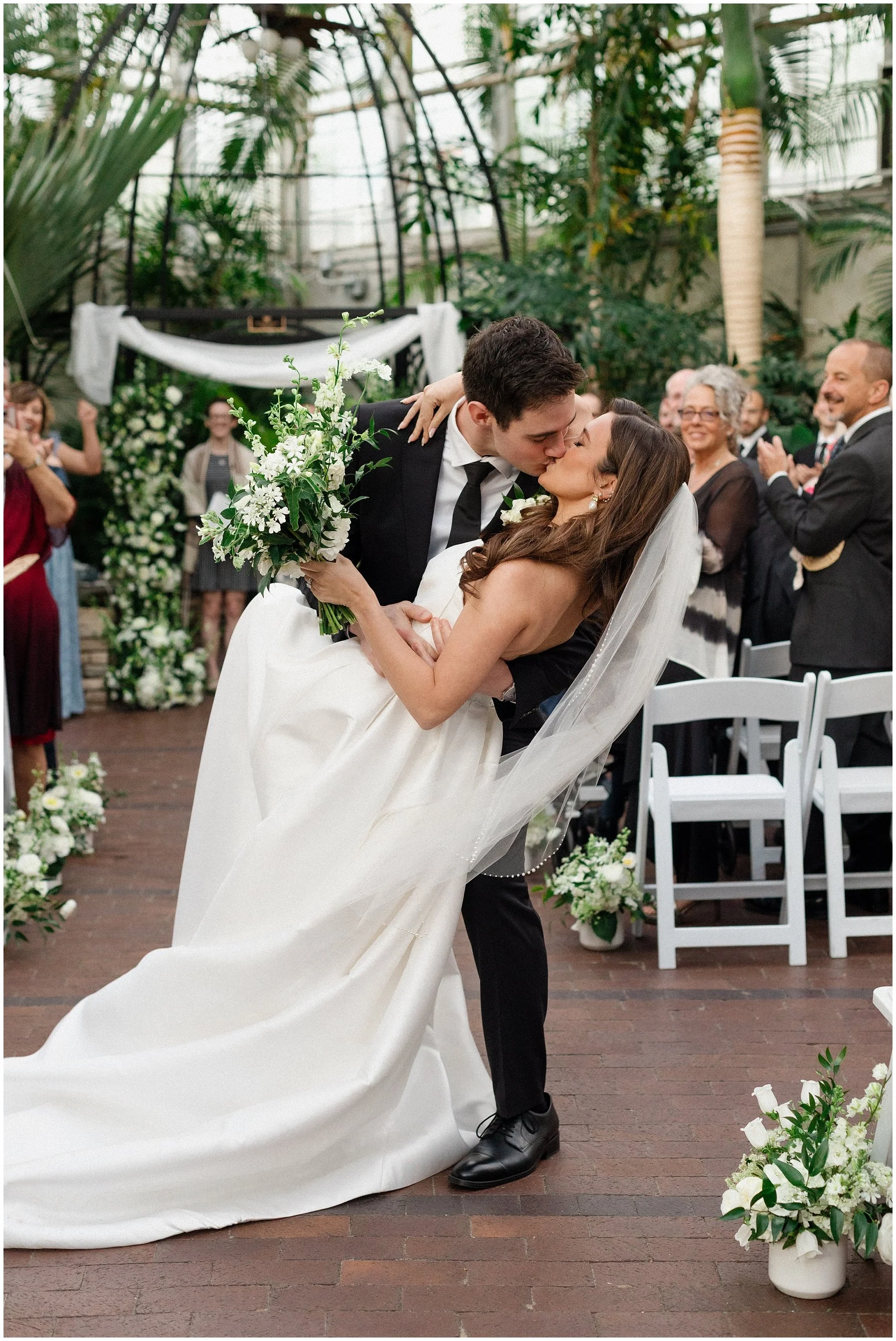 A newlywed couple shares a kiss at their wedding ceremony inside a greenhouse, with guests clapping and smiling in the background. The groom is in a black suit and the bride in a white wedding dress holding a bouquet of white flowers. Franklin Park C
