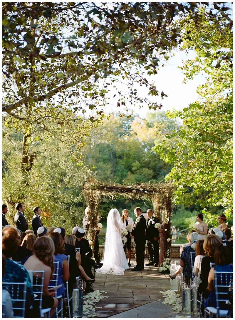 Outdoor wedding ceremony with a bride and groom exchanging vows under a decorated arch, surrounded by guests seated outdoors among trees.