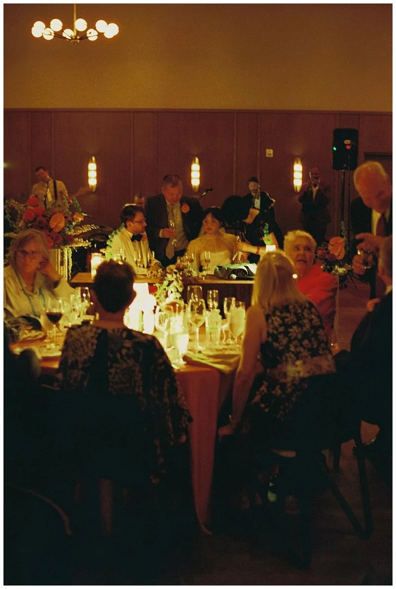 People socializing and dining at a formal event with candles, wine, and floral centerpieces, with a band playing in the background.