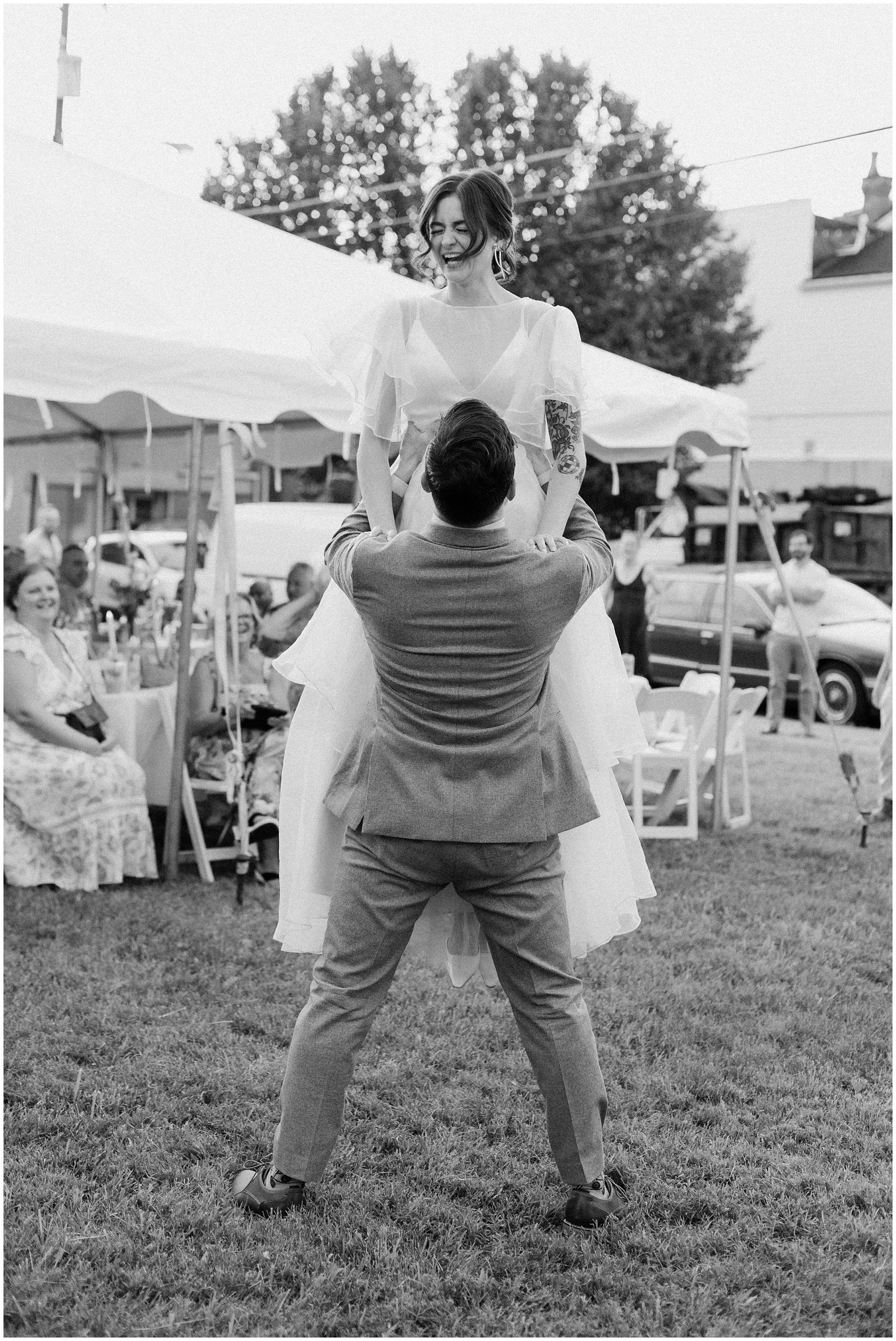 A person in a suit lifts a woman in a white dress and veil during a wedding celebration outdoors, with guests and cars in the background.