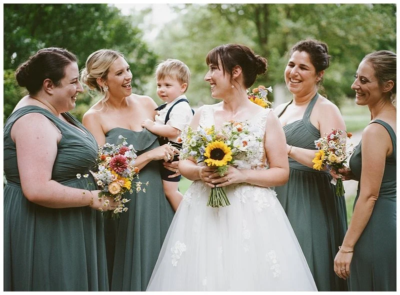 Bride in white dress holding sunflower bouquet surrounded by bridesmaids in teal dresses and a young boy in a suit during a wedding outdoor ceremony
