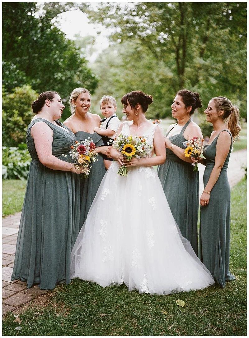 Bride in a white wedding gown holding a sunflower bouquet, surrounded by five women in matching gray dresses holding bouquets, and a young boy in a white shirt and suspenders holding her arm, outdoors with green trees.