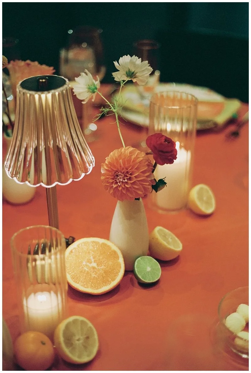 A table setting with a small floral arrangement, sliced citrus fruits, candles, and a pinkish-orange tablecloth.