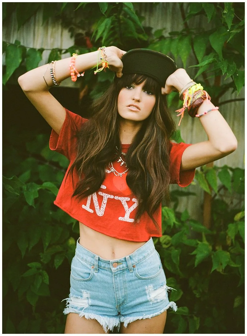 A young woman with long brown hair and bangs wearing a red crop top with the word 'NY' and denim shorts, posing outdoors with a black cap and layered bracelets, surrounded by green foliage.