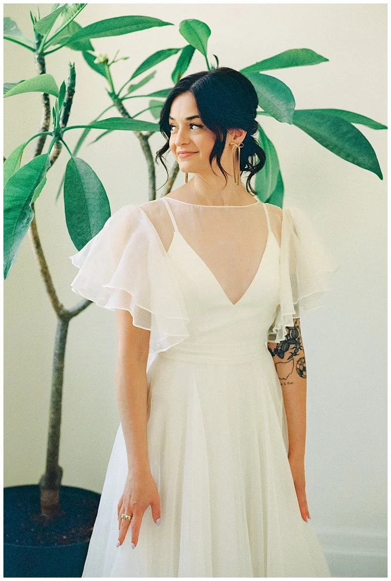 A woman in a white dress with ruffled sleeves standing next to a large green houseplant, smiling and looking to the side. Film Photography