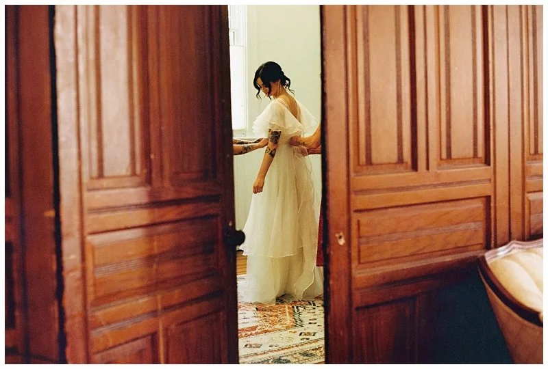 A woman in a white dress standing near a window, with someone behind her adjusting her dress, viewed through an open wooden door. Film Photography