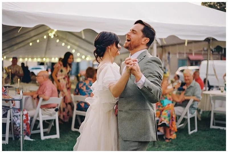 A bride and groom dancing at their outdoor wedding reception under a white tent with string lights, surrounded by seated guests.