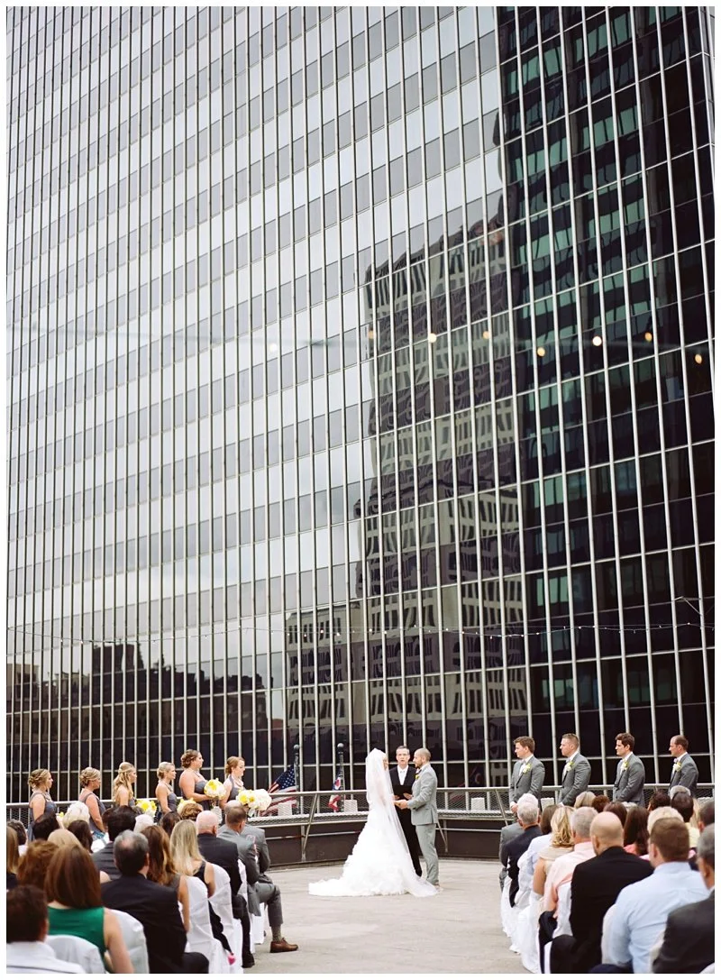 Outdoor wedding ceremony in front of a modern glass skyscraper with a reflection of another building, with the bride, groom, and wedding party standing in front of seated guests.