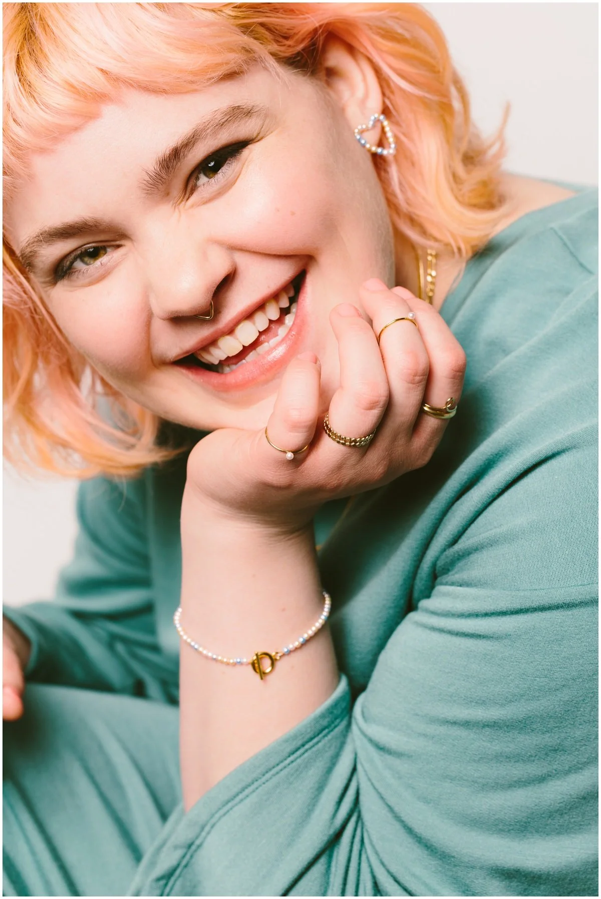 Close-up of a smiling woman with peach-colored hair, wearing multiple rings, earrings, a bracelet, and a light blue top, resting her chin on her hand. Portrait photography by Adam lowe photography Columbus Ohio studio fashion commercial branding. ADA