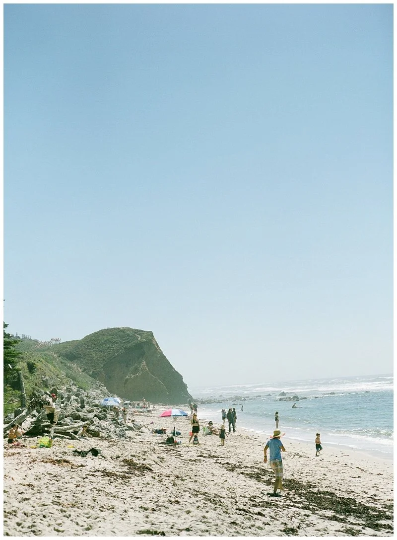 Beach scene with people sunbathing, umbrellas, and a rocky coastline under a clear sky.