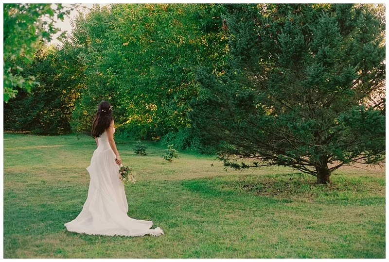 A bride in a white wedding gown holding a bouquet standing on grass next to a large evergreen tree with lush green foliage.