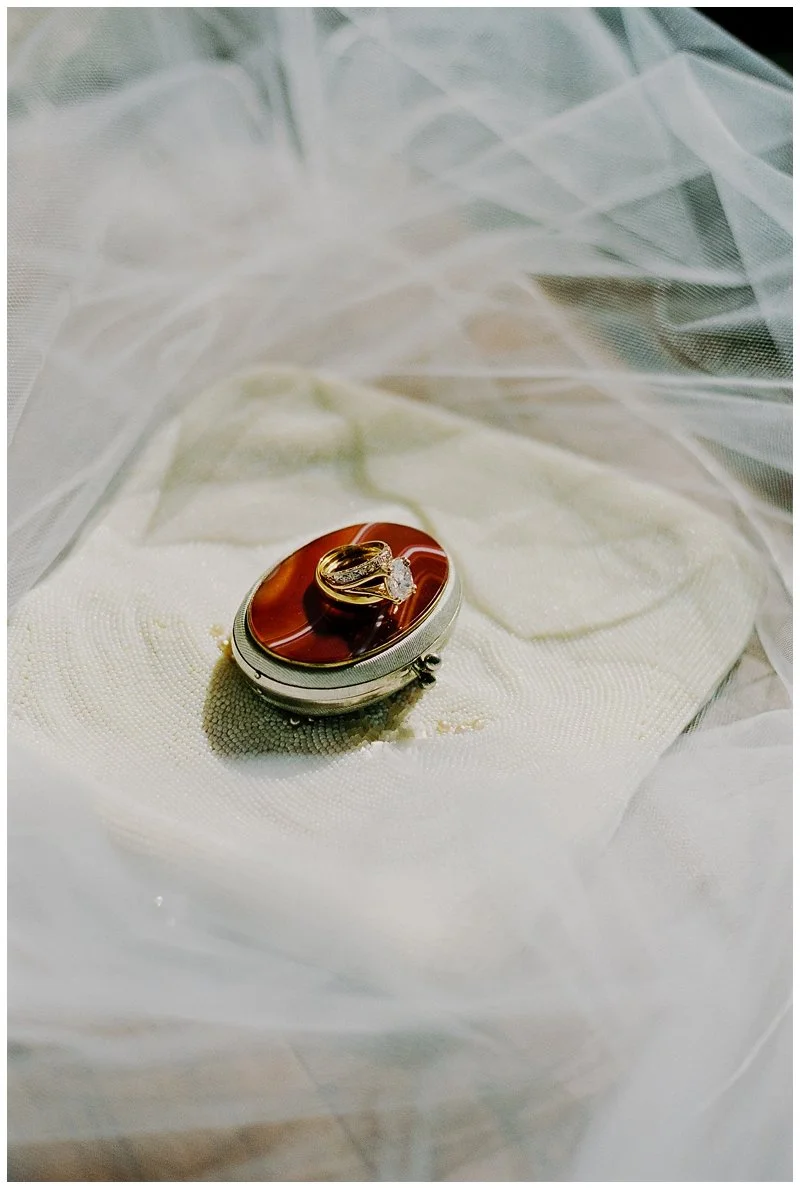 A ring with a large clear gemstone and gold band resting on an oval-shaped reddish-brown stone, placed on a white fabric surface.