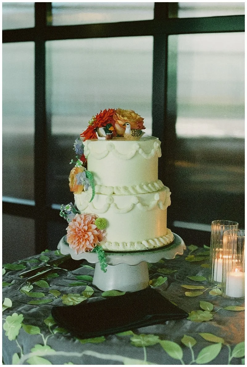 A two-tiered white wedding cake decorated with flowers, birds, and greenery, sitting on a white cake stand on a table with green leaf decoration and lit candles.