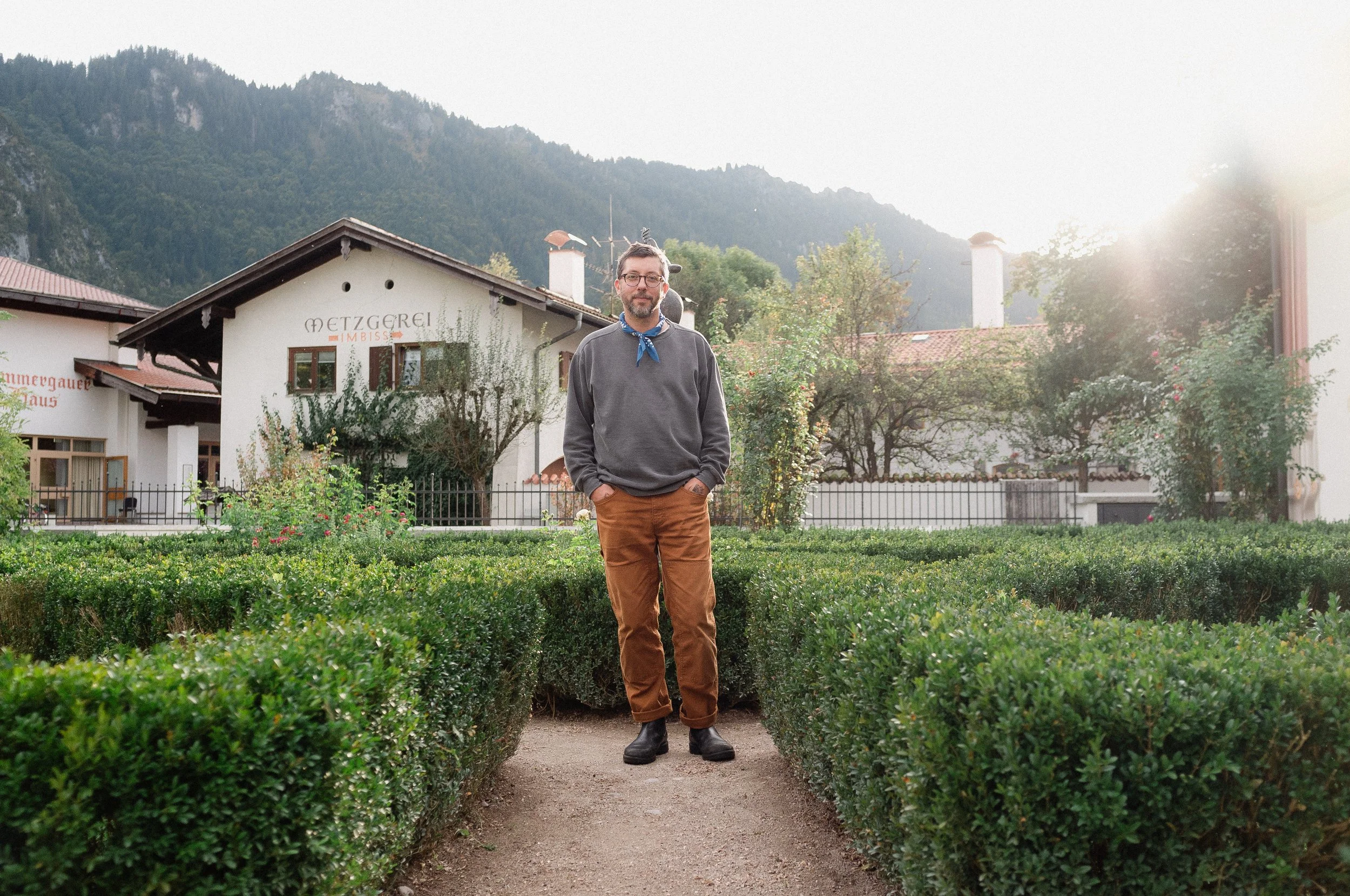 A man standing on a dirt path between trimmed hedges in front of a white building with brown window frames, mountains in the background, and the sun shining brightly from the right side of the image.
