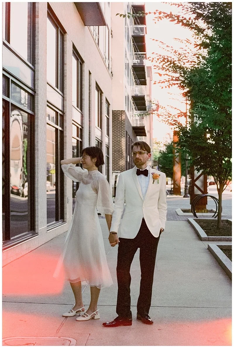 A couple dressed in wedding attire holding hands and walking on the sidewalk in an urban area during sunset or evening. The woman is wearing a white dress and heels, while the man is in a white tuxedo jacket, black pants, bow tie, and glasses.