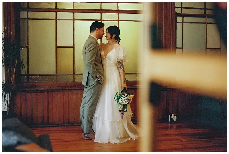 A bride and groom standing close to each other in a room with wooden floors and a wooden wall with window panels, looking at each other. The bride is holding a bouquet and has a tattoo sleeve on her left arm, while the groom is wearing a light gray s
