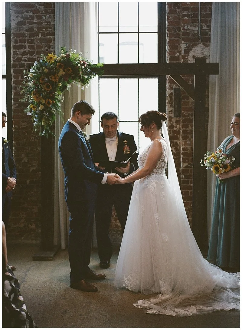 A bride and groom exchange vows in a rustic indoor wedding ceremony, with an officiant holding a book, in front of large industrial-style windows and a brick wall, with bridesmaids holding bouquets nearby.