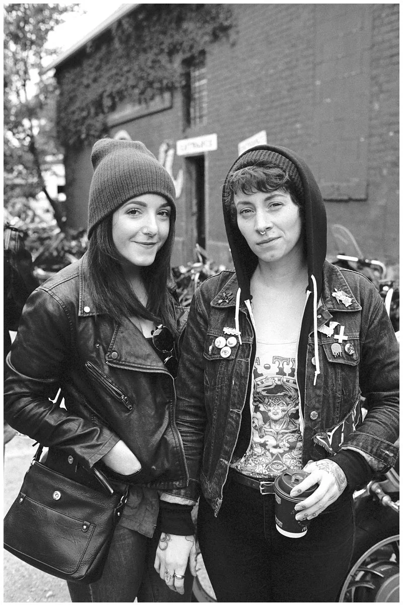 Two young women standing outdoors, dressed in black leather jackets, with one wearing a beanie and the other a hooded sweatshirt. They are smiling at the camera, with a brick building and bicycles in the background.