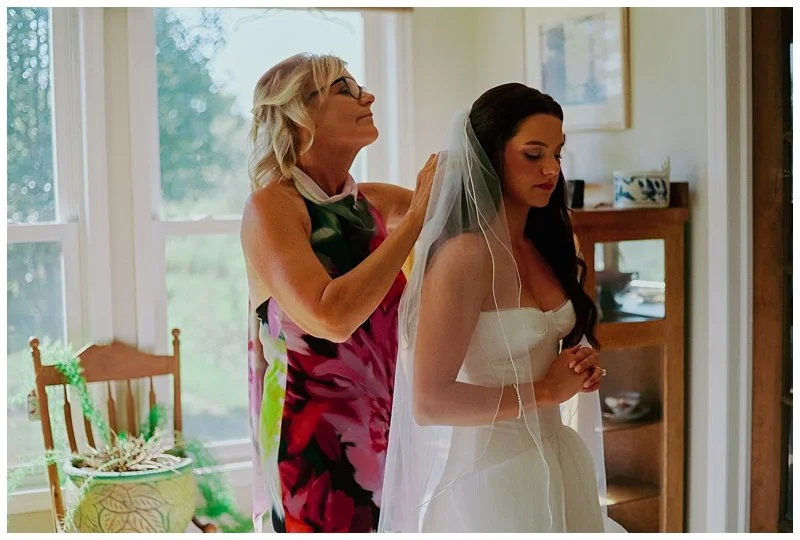 An older woman, likely the mother of a bride, helps adjust the bride's veil in a well-lit room with large windows. The bride is wearing a white dress and has her eyes closed, appearing contemplative. The room has a wooden chair with a pillow and some