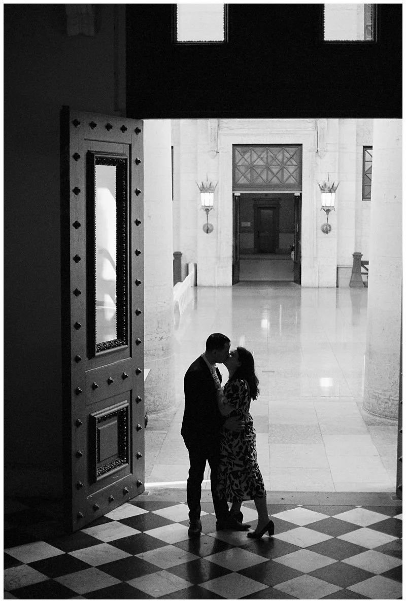 A silhouette of a couple kissing inside a grand building, with checkerboard flooring, large doors, and ornate wall sconces in the background.