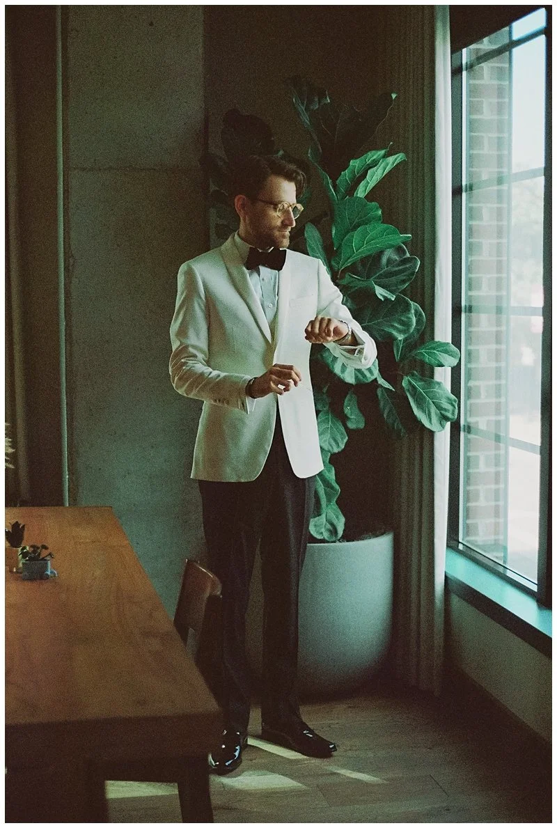A man in a white tuxedo jacket, black bow tie, and glasses is looking at his watch while standing indoors near a large window near a green potted plant.