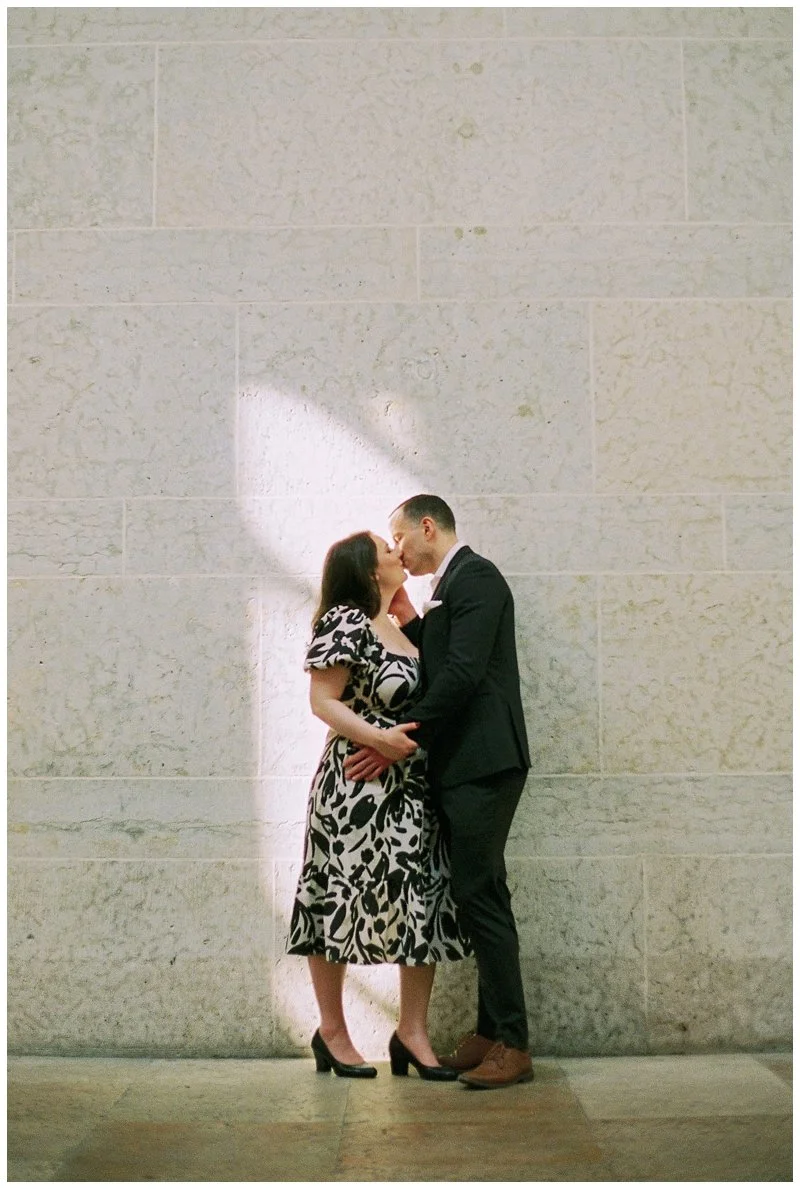 A couple is sharing a kiss against a textured wall, with the woman wearing a patterned dress and high heels, and the man in a dark suit and brown shoes.