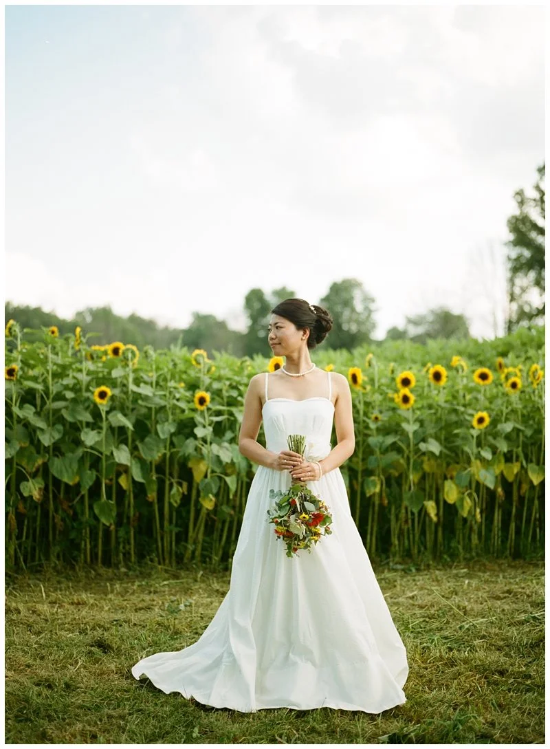 A woman in a white wedding dress holding a bouquet standing in a sunflower field.