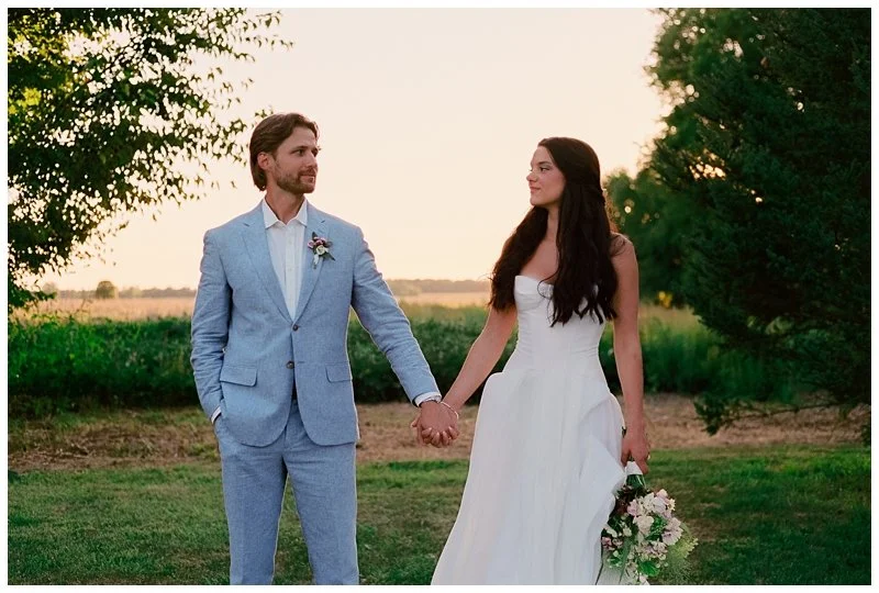 A bride and groom holding hands outdoors at sunset, with trees and open fields in the background.