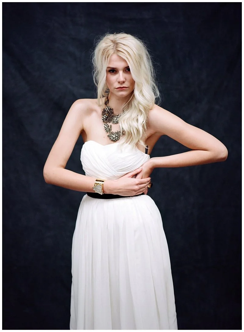 A woman with long, wavy platinum blonde hair, wearing a strapless white gown, black belt, and statement jewelry including a necklace, bracelet, and ring, standing against a dark background.
