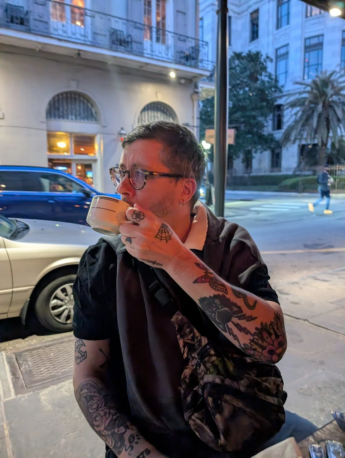 A man with tattoos, glasses, and earrings, sitting outside on a city street at dusk, drinking from a beige cup, with buildings, cars, and palm trees in the background.