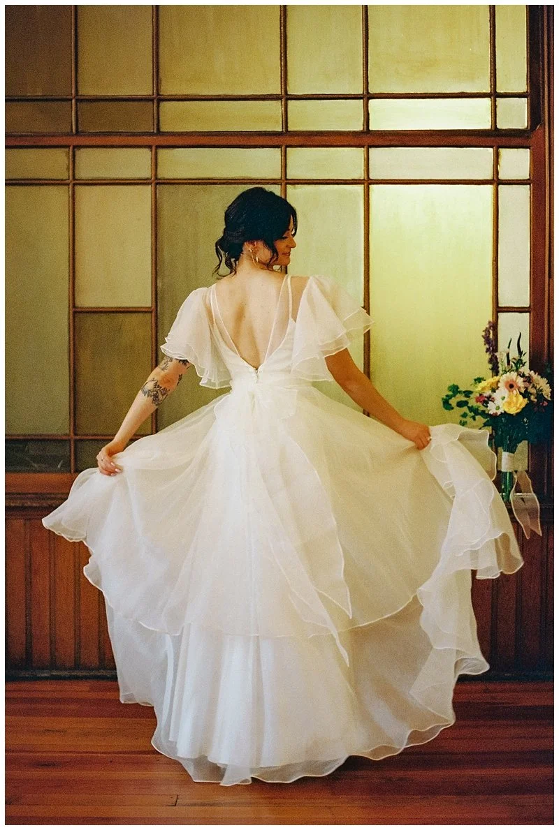 Woman in a white wedding dress with ruffled sleeves, standing in front of a wooden and glass wall, holding the dress and looking to the side, with a flower arrangement nearby. Film Photography