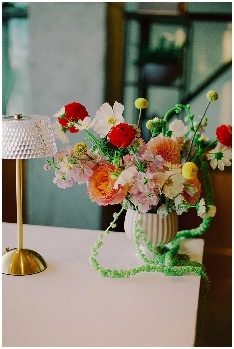 A white table with a decorative table lamp and a flower arrangement in a white ribbed vase. The flowers are a mix of pink, white, peach, red, and yellow blooms with green stems. There are also some light green fuzzy string-like decorations on the tab