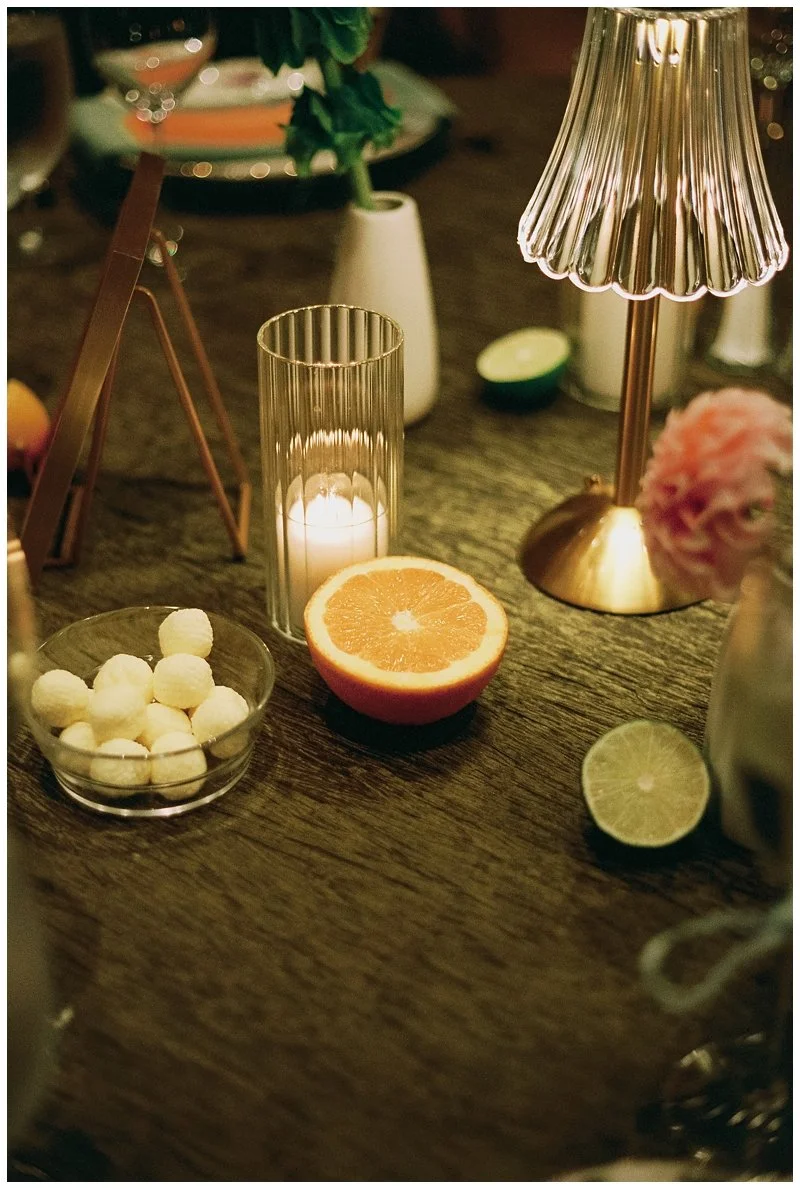 A cozy table setting with a lit candle, a halved orange and lime, a pink flower, and a small bowl of white balls on a wooden surface.
