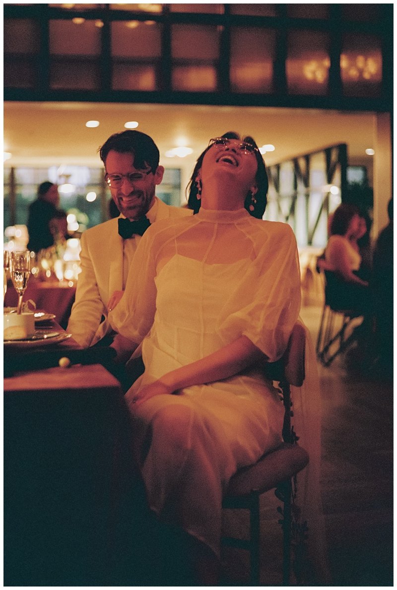 Two people laughing and enjoying themselves at a formal event or dinner in a warmly lit room. The man is wearing a white tuxedo with a black bow tie, and the woman is dressed in a pale yellow dress with short sleeves and earrings.