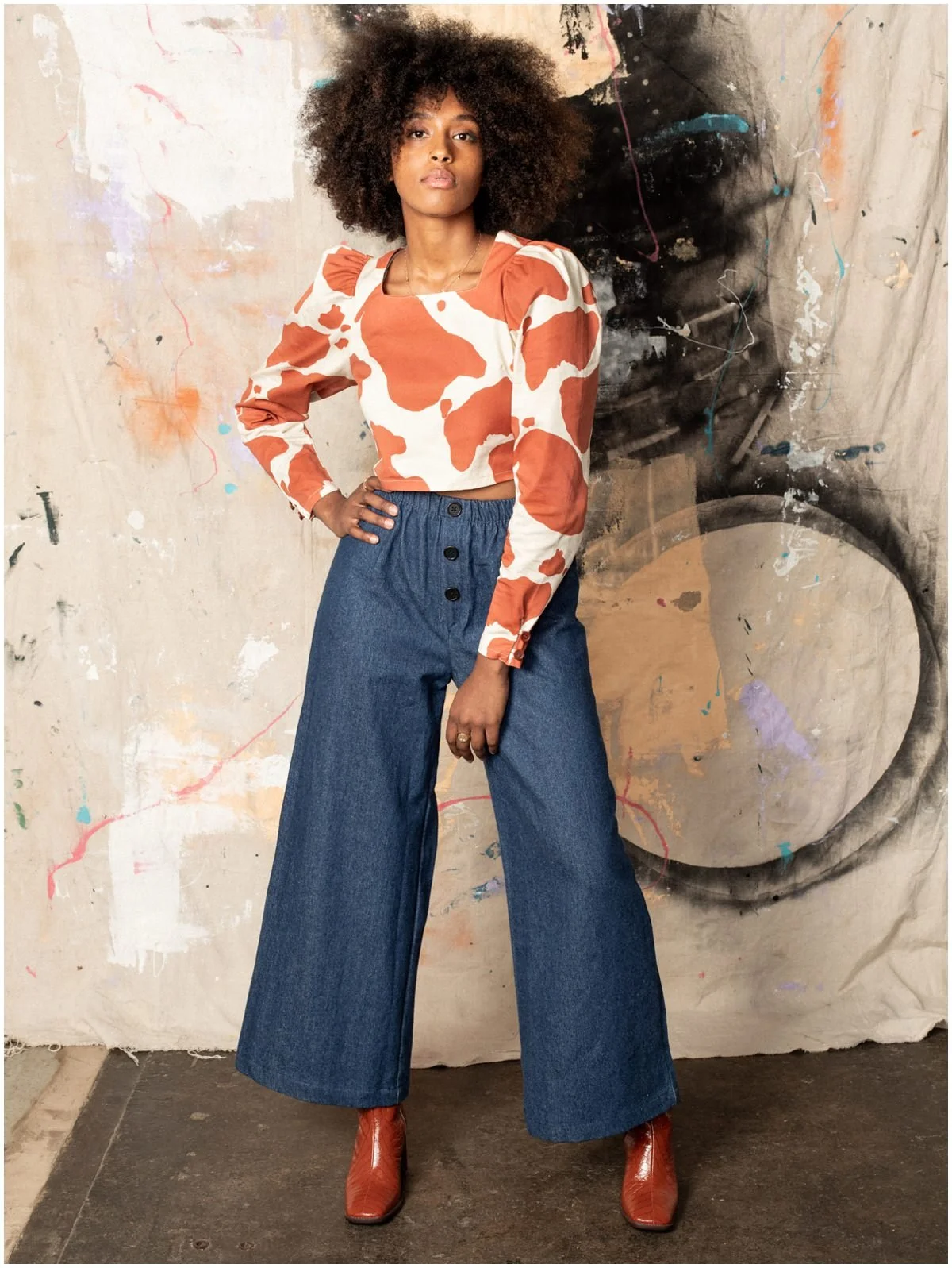 A woman with curly hair wearing a cow-print long sleeve top, wide-leg blue jeans, and orange ankle boots standing in front of an abstract art background.