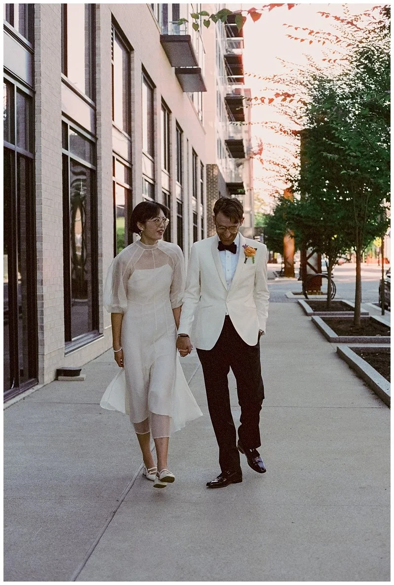 A newlywed couple walking hand-in-hand on a city sidewalk at sunset, dressed in formal wedding attire. The bride is wearing a white dress and glasses, and the groom is in a white tuxedo jacket, black pants, and a bow tie with a boutonniere. They are 