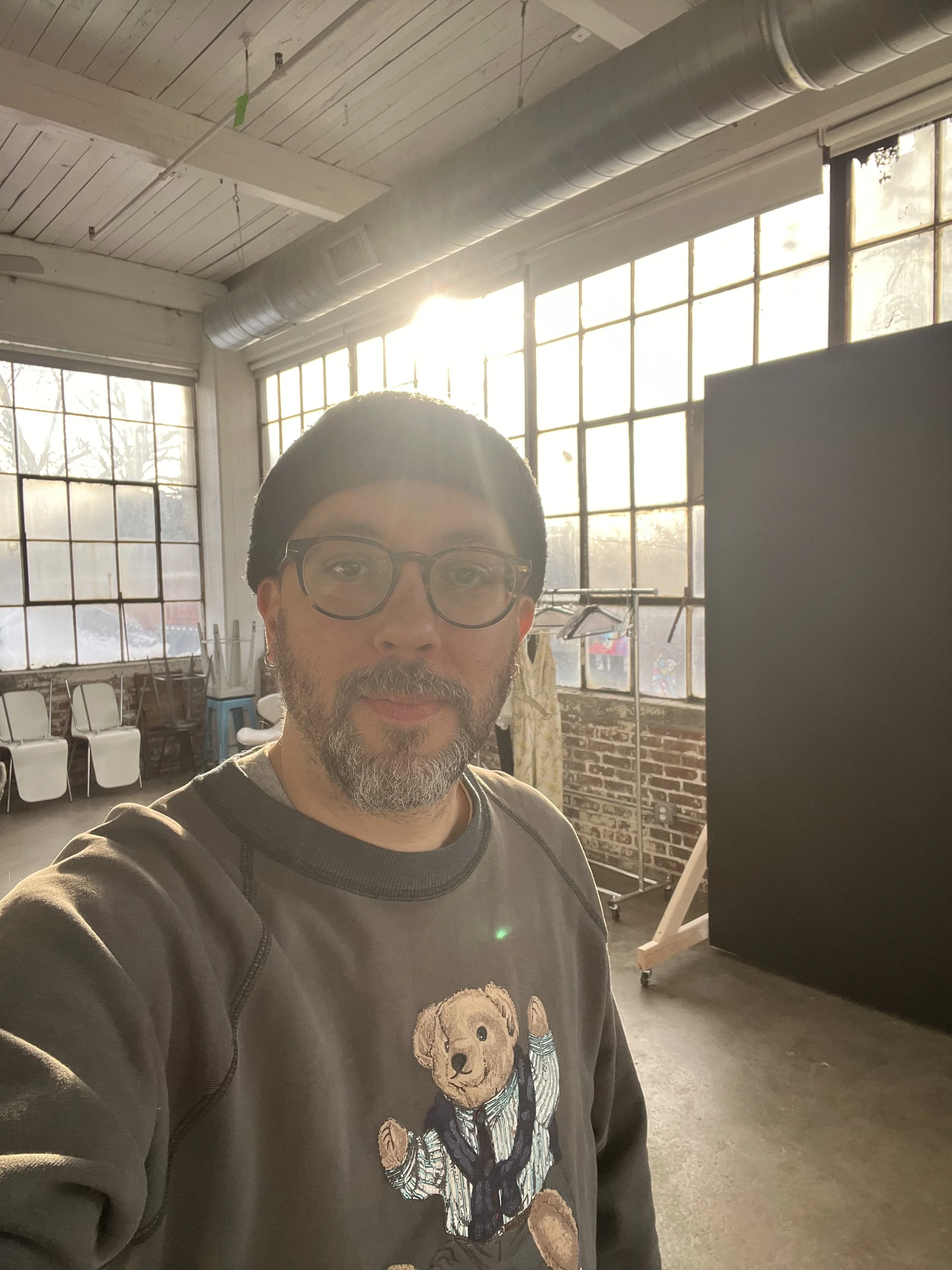 Man with glasses and a beard taking a selfie in a bright studio with large windows and a black backdrop.