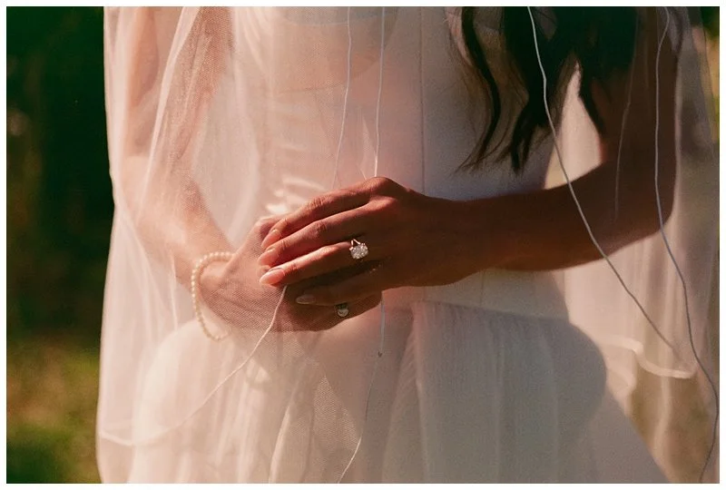 Close-up of a bride's hands with a wedding ring, outdoors in soft lighting, with sheer fabric and wavy hair in the background.