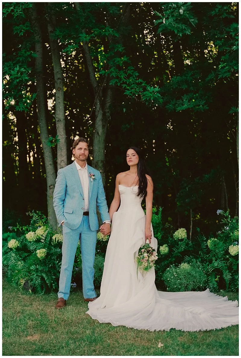 A bride and groom holding hands outdoors during their wedding. The bride wears a strapless white wedding gown and holds a bouquet, while the groom wears a light blue suit with a boutonnière. They stand on a grassy area in front of green foliage and t
