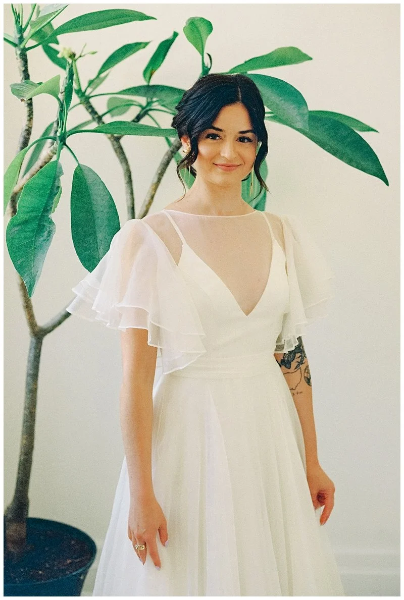A young woman wearing a white dress with sheer ruffled sleeves, standing indoors next to a large green potted plant, smiling at the camera. Film Photography