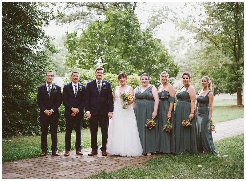 A wedding party standing outdoors on a brick pathway, with green trees and grass in the background. The group includes three men in dark suits and a woman in a white wedding dress holding a bouquet, along with four women in matching teal dresses hold