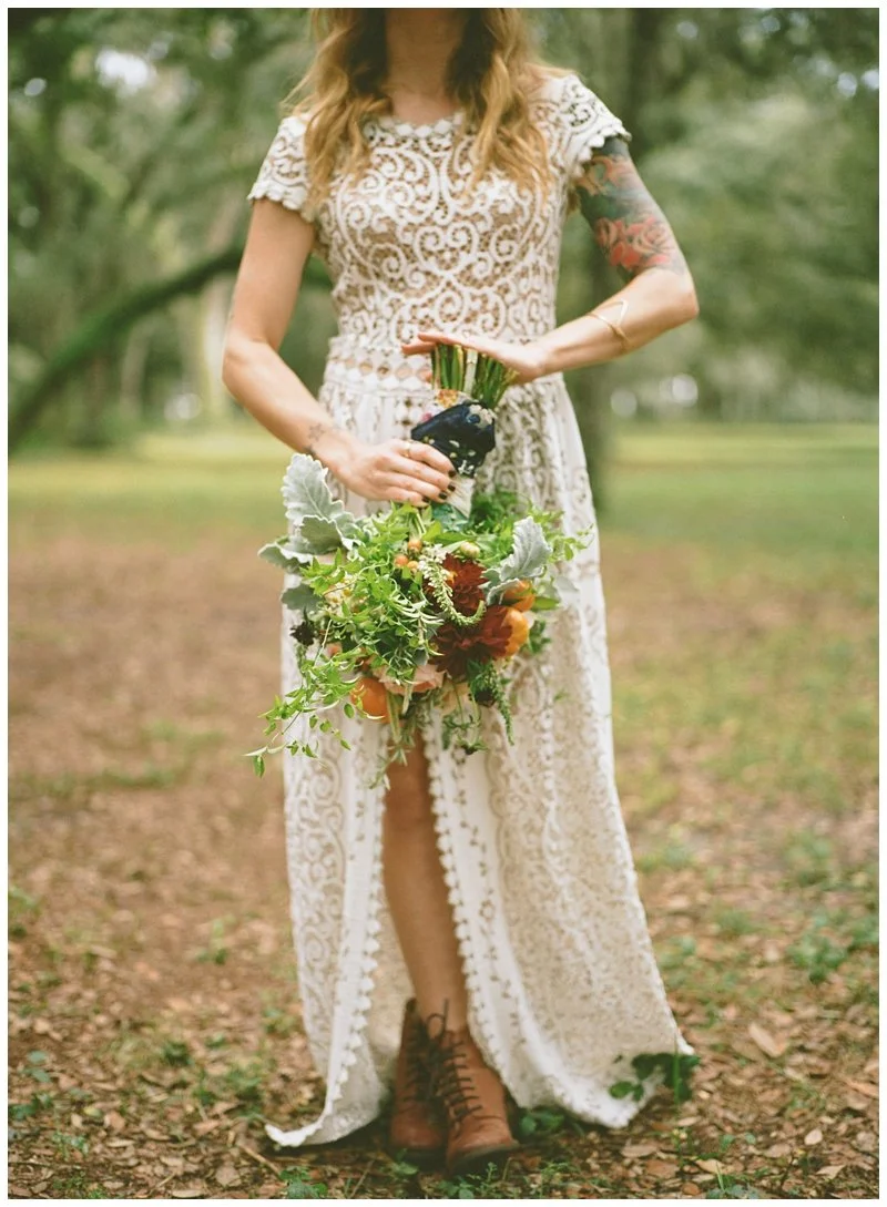 Woman in a lace dress holding a bouquet of flowers, standing outdoors on a dirt path in a park or forest setting.