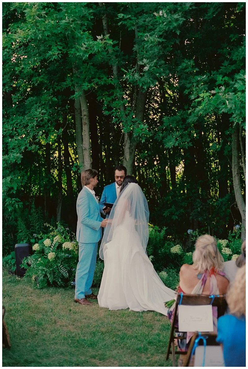 A couple getting married outdoors by a wooded area with officiant, bridesmaid, and guests in the background.